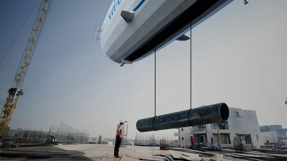 Construction worker overseeing a large pipe being lifted by crane at the Flying Whales airship production facility build site.