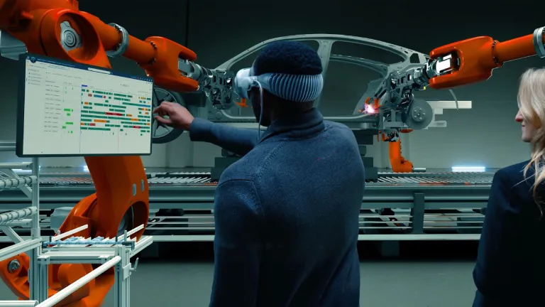 Sense Experiences Image Alt Text: Person wearing a mixed-reality headset reviews a 3DEXPERIENCE production-scheduling dashboard in an automated automotive factory, where robotic arms assemble a vehicle chassis.